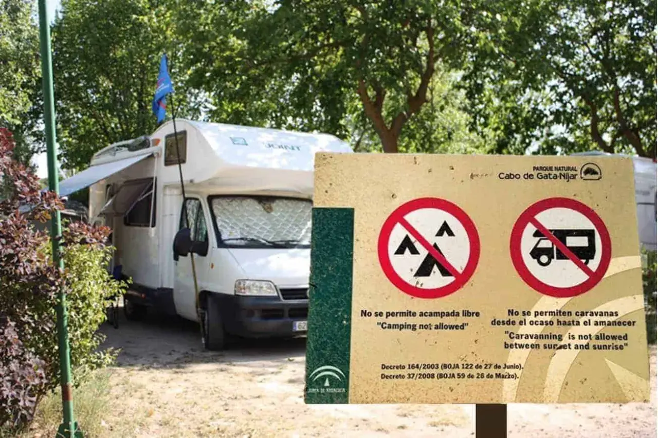 Una autocaravana blanca está aparcada junto a un cartel que prohíbe acampar y aparcar caravanas de noche.