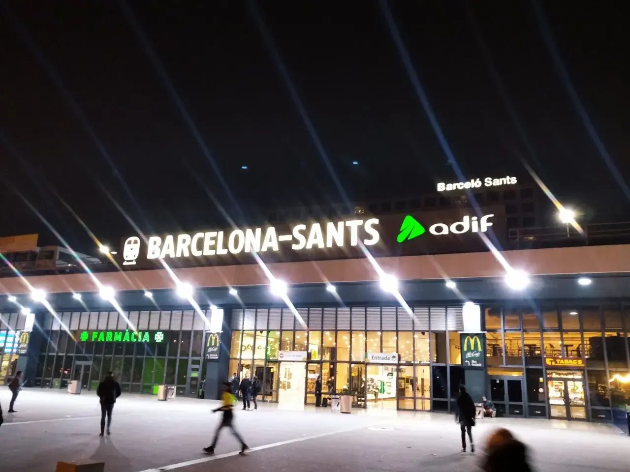 Noche en Barcelona Sants. Luces brillantes iluminan la fachada de la estación, con letreros de "Farmàcia" y "McDonald's".