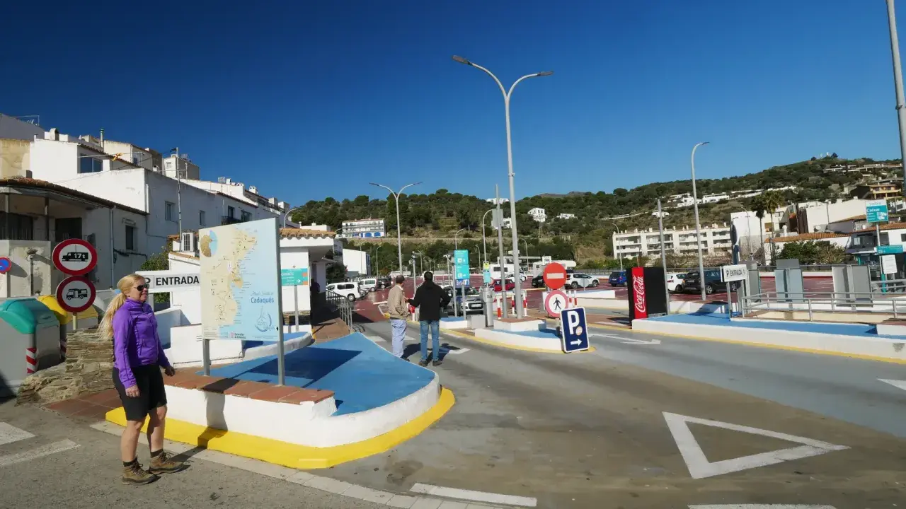 Entrada a parking gratuit Cadaqués. Una mujer rubia sonríe cerca de un mapa turístico. El cielo azul y las casas blancas dominan la escena.