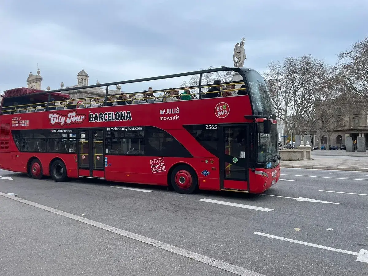 Autobús rojo de dos pisos "City Tour Barcelona" con turistas en la cubierta superior, ideal para el transporte en Barcelona para turistas.