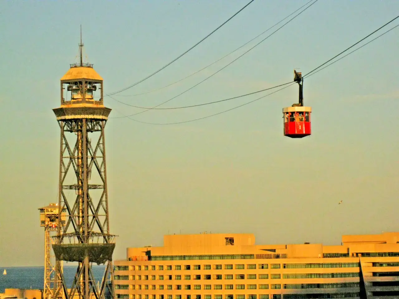 La cabina roja del montjuic seilbahn viaja sobre la ciudad, con una torre de transmisión y edificios modernos de fondo.