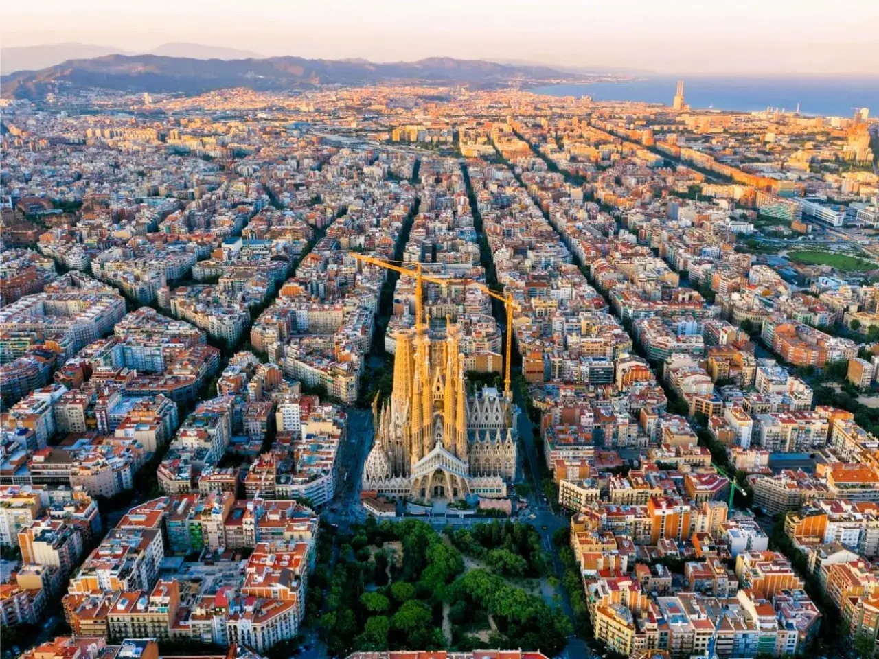 Vista aérea de Barcelona al atardecer, con la Sagrada Familia como punto focal. Las grúas de construcción rodean la basílica, símbolo de la ciudad.