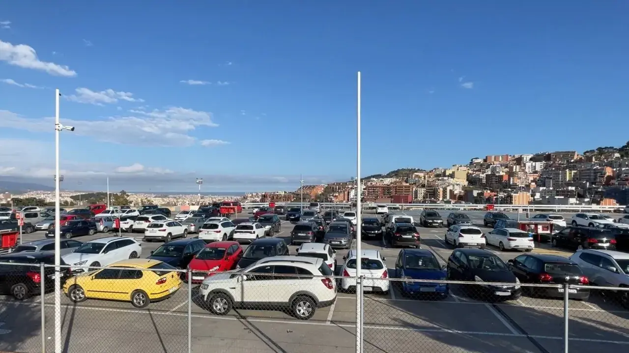 Amplio aparcamiento de coches en bsm Sant Genís, con la ciudad al fondo bajo un cielo azul.