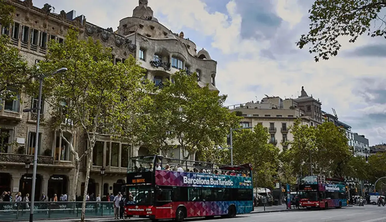 Autobuses tur&iacute;sticos en el Passeig de Gr&agrave;cia, cerca de la parada de metro. Edificios modernistas y &aacute;rboles verdes flanquean la calle.