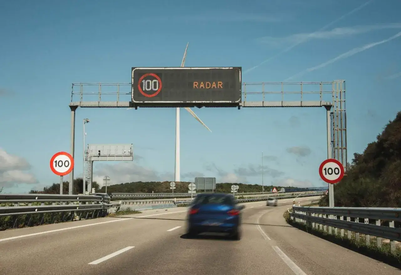 Un coche azul circula por una autopista bajo un cartel de radar de tramo que indica 100 km/h.