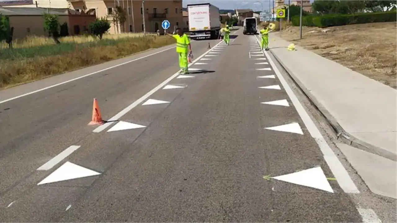 Trabajadores con chalecos reflectantes pintan nuevas marcas viales en la carretera. Conos naranjas y camiones al fondo.