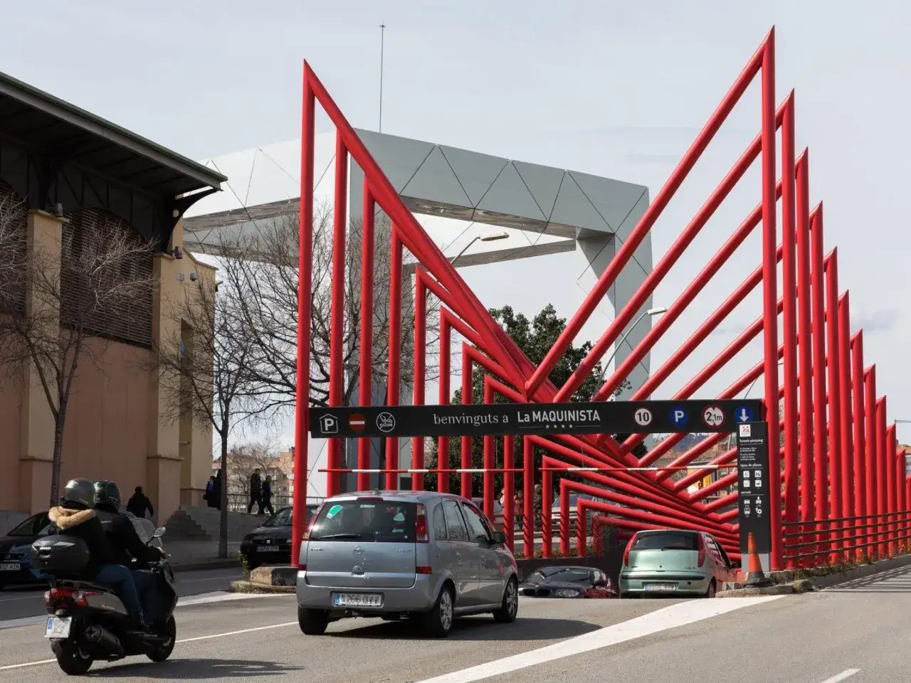 Entrada a la zona de la maquinista parking gratuit. Coches y moto circulan bajo una estructura roja y gris.