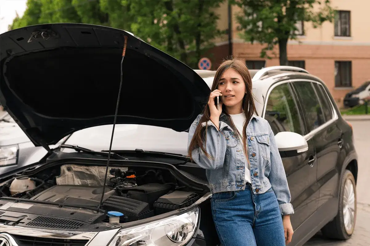 Mujer preocupada habla por teléfono junto a su coche con el capó abierto, quizás por tirones del coche.