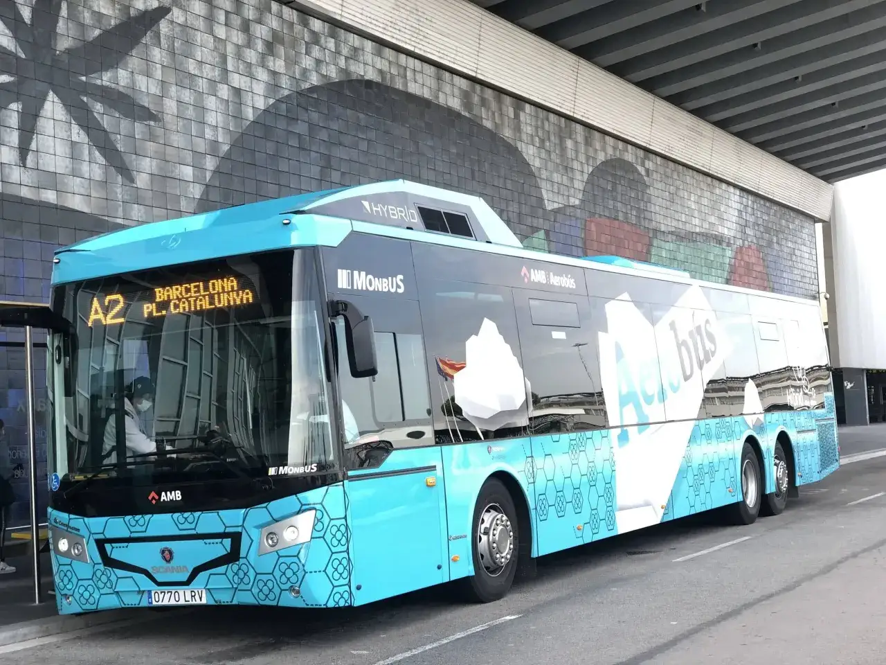 Autobús azul del Aerobús en Plaza Catalunya, Barcelona. El vehículo híbrido de Monbus se detiene bajo una pared con un diseño abstracto.