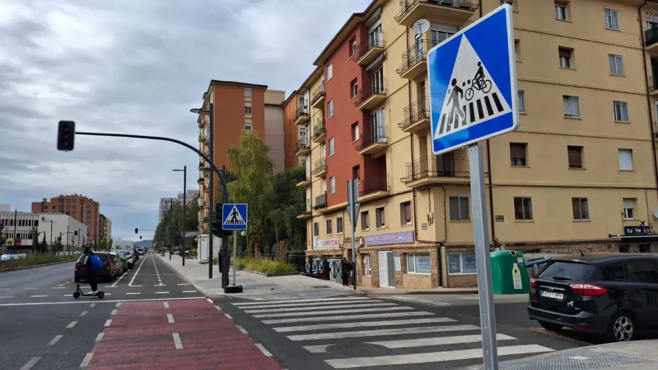 Señal de paso de peatones con un ciclista y un peatón, junto a edificios residenciales y una calle con carriles para bicicletas.