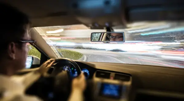 Conduciendo de noche bajo la lluvia, las luces de la carretera se difuminan. El conductor, con gafas, se refleja en el retrovisor, mostrando que es el ASR de un coche.